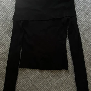 Svart offshoulder topp från H&M - Svart ribbad offshoulder topp från H&M i storlek M. Tröjan har långa ärmar och är tillverkad i ett mjukt material med 70% viskos. Snygg och enkel att styla med olika outfits, perfekt för dig som gillar minimalistisk stil.