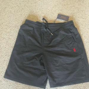 Svarta shorts Polo Ralph Lauren - Svarta shorts från Polo Ralph Lauren med klassisk röd logga broderad på benet. De har snörning i midjan och är tillverkade i ett mjukt bomullsmaterial. Perfekta för chill dagar eller när du vill ha en clean och stilren look. Skriv om du vill ha mer info