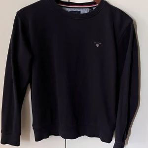 Marinblå sweatshirt från GANT - En stilren marinblå sweatshirt från GANT med rund halsringning och diskret logga broderad på bröstet. Tröjan har långa ärmar och är tillverkad i mjuk bomull, perfekt för en clean och klassisk look.