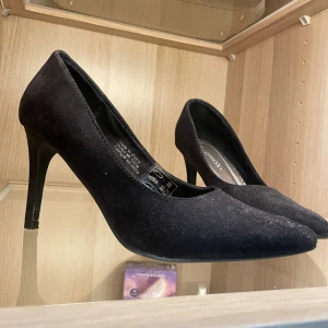Svarta pumps från Sprox, storlek 39 - Klassiska svarta pumps från Sprox i storlek 39. Skorna har spetsig tå och hög klack, tillverkade i mockaliknande material som ger en elegant look. Perfekta för dig som vill ha en stilren och tidlös sko med snygg siluett. Lite trötta i materialet