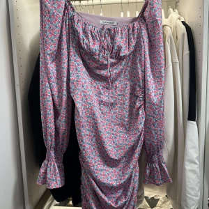 Blommig kortklänning från Adoore - Superfin kortklänning från Adoore med blommigt mönster i rosa, lila och blått. Klänningen har långa ärmar med volang vid ärmslut, snörning vid halsen och rynkade detaljer längs sidorna. Perfekt för dig som gillar färg och romantiska vibes!