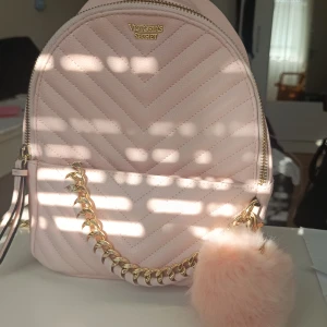 Ljusrosa ryggsäck från Victoria's Secret - Supersöt ljusrosa ryggsäck från Victoria's Secret med quiltad yta och guldfärgad kedjedetalj framtill. Väskan har dragkedja, randigt foder och en fluffig pompom som accessoar. Perfekt storlek för vardagens essentials och snygg att bära på ryggen. Jag köpte den från Victoria secret några år tillbaka och har knappt använt den. Den är helt slutsåld överallt och man kan inte hitta den typen längre. Finns en liten fläck längst ner på framsidan men inget man märker direkt. 