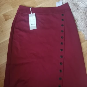 Vinröd kjol med knappar i sidenlook - Vinröd långkjol med snygga svarta knappar längs ena sidan och dragkedja bak. Kjolen har en stilren och rak passform i ett slätt material som ger en lyxig känsla. Perfekt för dig som vill ha en klassisk men trendig look.