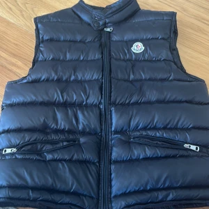 Svart dunväst från Moncler - Snygg svart dunväst från Moncler med quiltad design och hög krage. Västen har två dragkedjefickor framtill, diskret Moncler-logga på bröstet.