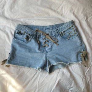 Ljusblå jeansshorts med nitar och snörning - Snygga ljusblå jeansshorts från Jin Lin med cool snörning framtill och råa kanter. Shortsen har guldiga nitar på fickorna och knytband i sidorna. Skriv för frågor!