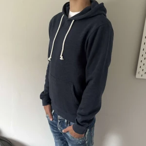 Mörkblå hoodie från Pier one - En stilren mörkblå hoodie med vita dragsnören och stor känguruficka framtill. Klassisk passform och mjukt material som känns skönt mot huden. Perfekt för dig som gillar en enkel och look.