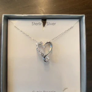 Halsband i silver med hjärthänge - Snyggt halsband i äkta silver med ett hjärtformat hänge dekorerat med gnistrande stenar av cubic zirkonia. Kedjan är tunn och elegant, perfekt för att ge en glittrig touch till din stil. Hjärtat har en modern, asymmetrisk design som känns både trendig och tidlös.
