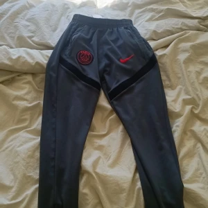 PSG x Nike grå mjukisbyxor - Grå mjukisbyxor från Nike med Paris Saint-Germain-logga och röd swoosh på vänster sida. Byxorna har svarta detaljer längs benen, elastisk midja och sidofickor. Perfekta för chill eller sport, med snygg och sportig vibe, de är äkta och jag har även koftan med till den ifall ni vill köpa den också, bara byxorna för 250kr och om ni ska ha koftan också så 350kr men 250kr styck för en av dem