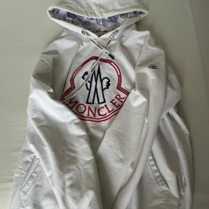 Vit Moncler hoodie med broderad logga - Säljer en vit hoodie från Moncler med stor broderad logga i rött och blått på bröstet. Tröjan har huva med snörning och mönstrat innerfoder samt en klassisk känguruficka. Moncler-märke på ärmen. Perfekt för dig som gillar stilrena och exklusiva plagg.
