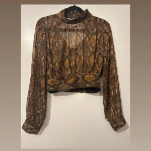 Brun croppad topp med ormmönster - Säljer en croppad långärmad topp med hög krage och ormmönster i bruna och svarta toner. Toppen har en liten öppning vid bröstet och är figurnära i modellen. Perfekt för vardag och fest.
