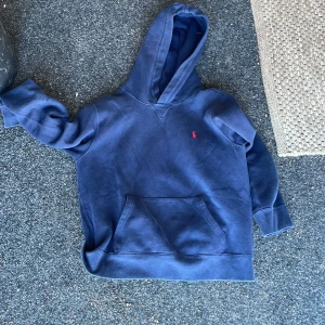 Mörkblå hoodie från Polo Ralph Lauren - Säljer en mörkblå hoodie från Polo Ralph Lauren med klassisk röd broderad logga på bröstet. Tröjan har huva, känguruficka och långa ärmar. Perfekt för dig som gillar en enkel och stilren look. Slitningar på höger arm