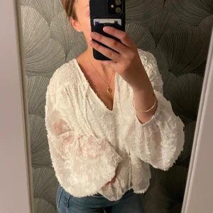 Vit blus med volymärmar från Gina Tricot! - Supersöt vit blus från Gina Tricot med v-ringning och voluminösa, transparenta ärmar. Blusen har ett broderat blommönster och lätt struktur i tyget som ger en romantisk vibe. Perfekt att styla med jeans eller kjol för en trendig look. Använd 1 gång!