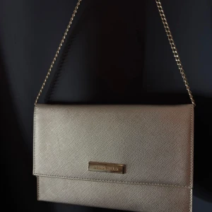 Guldig/champange kuvertväska från Anna Field - Stilren kuvertväska i guld/champange från Anna Field med elegant kedjerem. Väskan har en slät, lätt skimrande yta och ett diskret metallmärke framtill. Perfekt storlek för det viktigaste och med flera fack inuti. Snygg accessoar som lyfter din outfit. Kedjan går även att koppla loss, se sista bild. Inköpt från Zalando och endast använd en gång så den är som ny! 