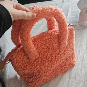 Fluffig orange handväska med dragkedja - Supergullig fluffig handväska i orange med mjukt teddy-material och rundade handtag. Väskan har dragkedja upptill och ett rymligt fack inuti. Perfekt för dig som vill sticka ut med en färgglad accessoar.