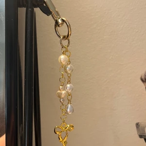 Guldig nyckelknippa med rosett - Unik nyckelknippa med guldpläterad kedja och oregelbundna vita pärlor i olika former. Längst ner hänger en söt liten rosett i guld. Perfekt accessoar för att lyfta din väskas look med en touch av elegans och lekfullhet. Handgjort!💓