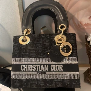 Christian Dior svart handväska - Snygg svart handväska från Christian Dior med tydlig logga framtill och guldfärgade metalldetaljer. Väskan har ett diskret mönster i tyget och robusta handtag i skinn. Perfekt accessoar för att lyfta din outfit.