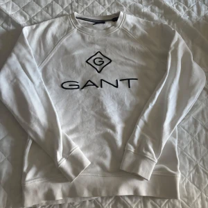 Vit sweatshirt från GANT med logga - Snygg vit sweatshirt från GANT med svart broderad logga på bröstet. Klassisk rund halsringning och långa ärmar. Tillverkad i mjuk bomull som är skön att ha på sig. Perfekt för en clean och stilren look.