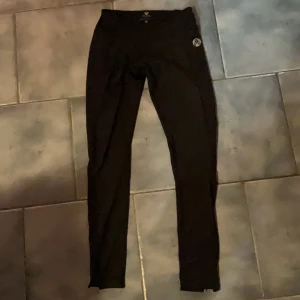 Svarta träningsleggings från Laplandic - Svarta träningsleggings från Laplandic med tajt passform. Byxorna har röda dragkedjor vid benslut och bakficka, samt hög midja. Perfekta för träning och aktiva dagar. Materialet är stretchigt och andas bra.