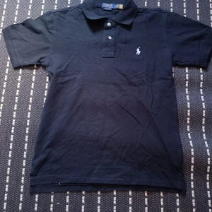 Svart pikétröja Polo Ralph Lauren M - Klassisk svart pikétröja från Polo Ralph Lauren i bomull. Tröjan har korta ärmar, krage med knappar och den ikoniska broderade loggan på bröstet. stor i storleken