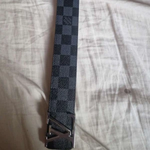 Louis Vuitton rutigt svart bälte - Snyggt svart bälte från Louis Vuitton med klassiskt rutigt mönster och ikoniskt LV-spänne i silver. Bältet är gjort i skinn och har en stilren design som passar till många outfits.