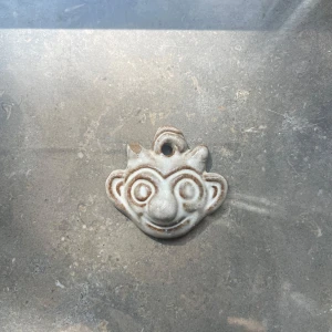 Keramiskt halsband trollansikte - Unikt handgjort halsbandshänge i keramik format som ett lekfullt trollansikte. Hängsmycket är beige med bruna detaljer och har ett hål upptill för kedja eller snöre.