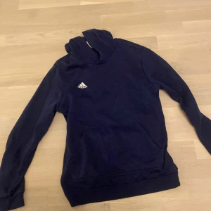 Mörkblå Adidas hoodie med huva - Säljer en mörkblå hoodie från Adidas med klassisk vit logga på bröstet. Tröjan har huva, känguruficka och långa ärmar. 