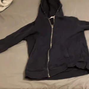 Svart hoodie med dragkedja från 157 - Svart hoodie från 157 med hel dragkedja och huva. Klassisk modell med två fickor framtill och ribbade muddar. Perfekt för chill dagar eller när du vill ha en enkel och stilren look. Mjuk insida och normal passform.