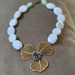 Blommigt halsband från Hultquist - Unikt halsband från Hultquist med stora vita pärlor och en stor blomma i guldtonad metall som hänge. Blommans mitt är dekorerad med små ljusblå pärlor. Perfekt statement-piece för dig som vill sticka ut.
