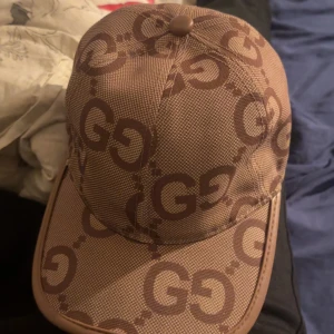 Gucci beige keps med GG-mönster - Snygg beige keps från Gucci med det klassiska GG-logomönstret i brunt över hela kepsen. Kepsen har böjd skärm och detaljer i läder-look längs kanten. Perfekt för dig som vill sticka ut med en lyxig accessoar.