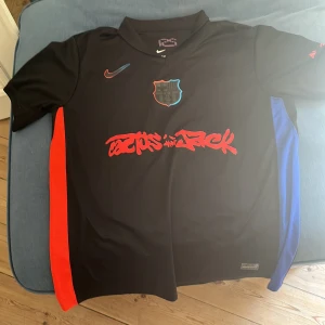 FC Barcelona x Nike Travis Scott tröja - Säljer en svart FC Barcelona x Nike fotbollströja med Travis Scott Cactus Jack-grafik i rött över bröstet. Tröjan har klubbmärke med blå och röd kant, Nike-logga i rött och blått samt röda och blå sidopaneler. Tillverkad i lätt och ventilerande material.