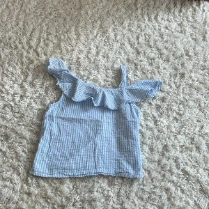 Blåvit randig linnetopp H&M volang - Supersöt blåvit randig linnetopp från H&M med volangdetalj längs halsringningen och en axelbandslös design på ena sidan. Perfekt för varma dagar med sin lätta känsla och somriga look. Sval och luftig, passar perfekt till shorts eller kjol.