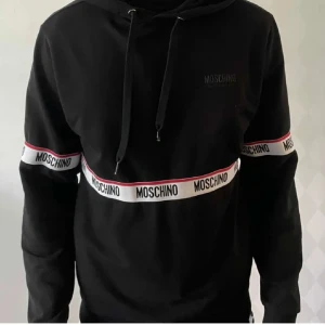 Svart Moschino hoodie med logoband - Svart hoodie från Moschino med vita och röda logoband runt bröstet och ärmarna. Klassisk huva med snörning och lång ärm. Snygg streetstil och perfekt för dig som gillar statement-plagg. Materialet känns mjukt och skönt, troligen bomull eller polyester.