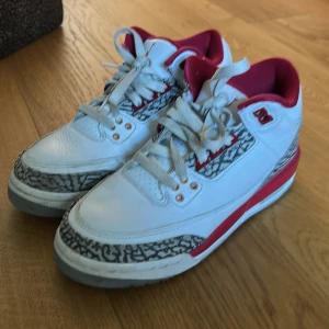 Nike Air Jordan 3 Cardinal Red - Säljer ett par Nike Air Jordan 3 Retro med vit läderöverdel, röda detaljer och ikoniskt elefantmönster i grått. Skorna har snörning, rund tå och klassisk Jumpman-logga på hälen. Perfekta för dig som vill sticka ut med en tidlös sneaker.