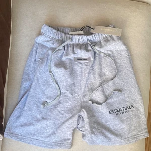 Grå shorts Essentials Fear of God - Snygga grå shorts från Essentials Fear of God med elastisk midja och snörning. Mjukt bomullsmaterial, relaxed fit och diskret logga framtill. Perfekta för chill dagar eller streetwear-stil. Fickor på sidorna och en bakficka för extra funktion. Helt oanvänd men plastpåsen är lite sliten. Taggen är sönder men det spelar inte så mycket roll.