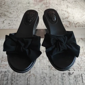 Svarta sandaler med rosett från QOG - Stilrena svarta sandaler från OOG generation med bred rem och stor rosett framtill. Platt sula med dekorativa sömmar längs kanten. Perfekta för varma dagar när du vill ha en enkel men snygg look.