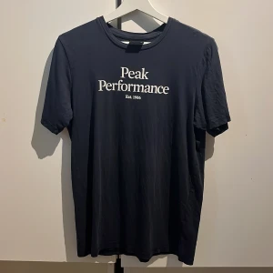 Mörkblå Peak Performance t-shirt - Snygg mörkblå t-shirt från Peak Performance med vit logga och text på bröstet. Klassisk rund halsringning och korta ärmar. Tillverkad i mjuk bomull som känns skön mot huden. Perfekt för en clean och sportig look.