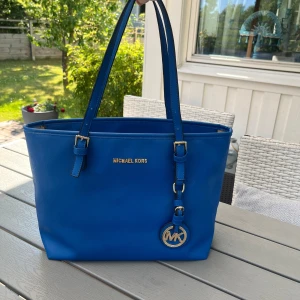 Blå Michael Kors axelväska i skinn - Snygg blå axelväska från Michael Kors i skinn med guldiga detaljer och MK-logga. Väskan har justerbara axelband, dragkedja upptill och ett rymligt innerfack med beige MK-mönstrat foder. Perfekt för dig som vill ha en stilren och praktisk väska.