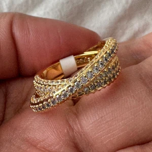 By Jolima - Donna twist ring, 14K guld med stenar - Detta var en present till en vän, men den var tyvärr för stor. Helt oanvänd i storlek 17 mm. Snygg twistad guldpläterad ring från by Jolima med glittrande stenar. Ringen har en cool korsad design som verkligen sticker ut och ger en lyxig känsla. Så fin!  Nypris, 499 SEK