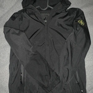 Svart vindjacka från Stone Island - Svart vindjacka från Stone Island med ikonisk patch på ärmen. Jackan har huva, dragkedja framtill och två sidofickor med dragkedja. Tillverkad i lätt polyester med slät insida. Perfekt för dig som vill ha en clean och stilren look.