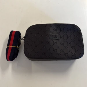 Svart axelväska från Gucci i skinn - Säljer en svart axelväska från Gucci i skinn med präglat GG-mönster över hela väskan. Väskan har en avtagbar axelrem i tyg med röd och marinblå rand. Stängs med dragkedja och har en stilren, rektangulär form. Perfekt för dig som gillar lyxiga detaljer.