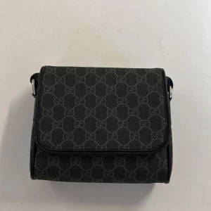 Svart axelväska från Gucci - Snygg svart axelväska från Gucci med klassiskt GG-mönster över hela väskan. Väskan har en justerbar axelrem i rött och marinblått, samt detaljer i skinn. Perfekt storlek för det viktigaste och en ikonisk accessoar till din outfit.