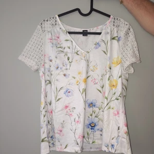 Blus - Superfin vit blus med blommigt mönster i pastellfärger som blått, rosa och gult. Blusen har v-ringning och korta ärmar i spets som ger en romantisk vibe. Perfekt för vår och sommar när du vill ha något luftigt och sött. Helt ny
