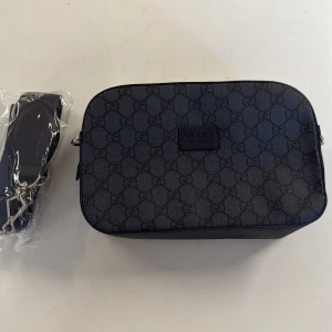Svart axelväska från Gucci med GG-mönster - Snygg svart axelväska från Gucci med klassiskt GG-mönster över hela väskan. Rektangulär form, justerbar axelrem och dragkedja upptill. Tillverkad i slitstark canvas med detaljer i skinn. Perfekt för dig som vill ha en stilren och lyxig väska med ikonisk design.