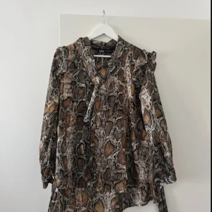 Blus med ormmönster - Snygg blus från Zara med trendigt ormmönster i bruna, svarta och beige toner. Blusen har långa ärmar, volangdetaljer vid axlarna och knytband vid halsen. Perfekt för dig som vill sticka ut med en edgy look.
