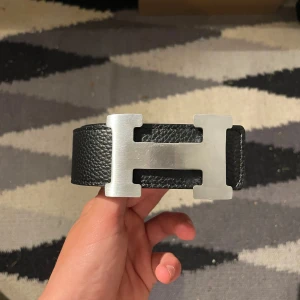skärp med H-spänne - Säljer ett bälte i äkta skinn med ikoniskt H-spänne i silverfärgad metall. Bältet är vändbart med ena sidan i svart och andra i mörkbrunt, båda med snygg struktur och vita kontrastsömmar. Perfekt accessoar för att lyfta din outfit.