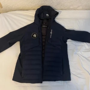 Sail Racing blå dunjacka med huva - Snygg mörkblå Sail Racing JR Antarctica Hybrid dunjacka med huva och coola detaljer som patch på ärmen och logga på bröstet. Jackan är quiltad framtill och har dragkedja. Perfekt för kalla dagar och har en sportig vibe.