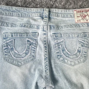 True Religion ljusa bootcut jeans - Säljer ett par ljusblå jeans från True Religion, modell CARRII Low Rise Flare. Jeansen har klassiska broderade bakfickor och bootcut-ben. Tillverkade i jeansmaterial med dragkedja och knapp framtill. Perfekta för dig som gillar retrovibbar och snygga detaljer.