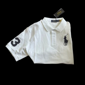 Ralph Lauren piké - Helt ny Vit pikétröja från Polo Ralph Lauren med klassisk krage, korta ärmar och svart broderad logga på bröstet. Snygg detalj med en svart siffra 3 på ena ärmen. Tillverkad i mjuk bomull som känns skön mot huden. Perfekt för en clean och sportig look.  Axelbredd 48cm. 