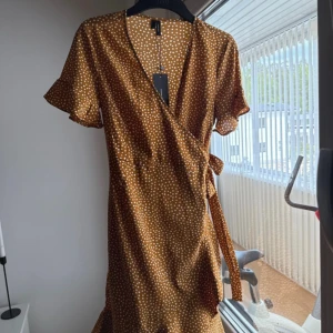 Prickig omlottklänning från Vero Moda - Söt kort omlottklänning från Vero Moda i varm brun färg med vita prickar. Klänningen har korta ärmar med lätt volang, knyt i midjan och v-ringad hals. Perfekt för sommarens alla tillfällen och ger en fräsch och trendig look.