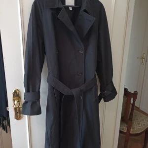 Svart trenchcoat med bälte - Stilren svart trenchcoat med klassisk krage, knappar framtill och matchande bälte i midjan. Jackan har långa ärmar med snygga spänndetaljer vid handlederna. Perfekt för dig som vill ha en tidlös och clean look.
